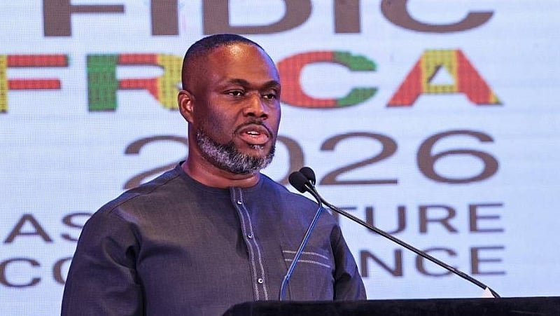 Man in a dark shirt speaking at a podium with a microphone event backdrop showing Africa branding and the year 2026