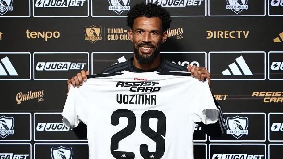 Male soccer player stands in front of a sponsor backdrop holding up a white jersey with the name VOZINHA and the number 29