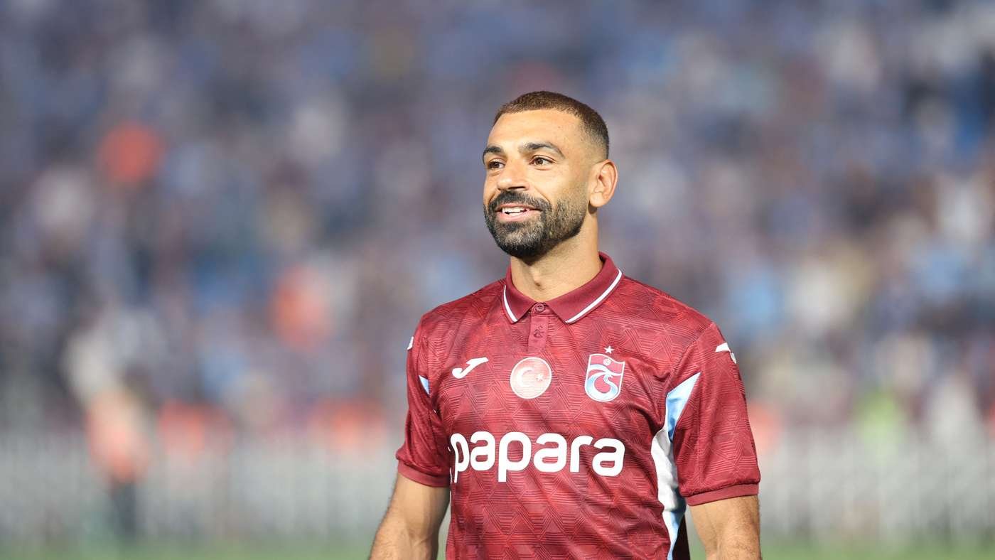 Male soccer player in a maroon jersey with the Papara sponsor smiling on the pitch with a blurred stadium crowd behind him