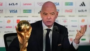 Bald man in a suit at a press conference with a FIFA badge gesturing with one hand a World Cup trophy on the table beside him
