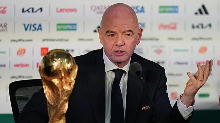 Bald man in a suit at a press conference with a FIFA badge gesturing with one hand a World Cup trophy on the table beside him