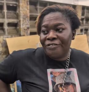 African woman with short hair wearing a black graphic T shirt and chain necklace speaking outdoors with wooden pallets in the background