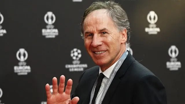 Smiling man in a dark suit waving at a step and repeat backdrop with logos behind him