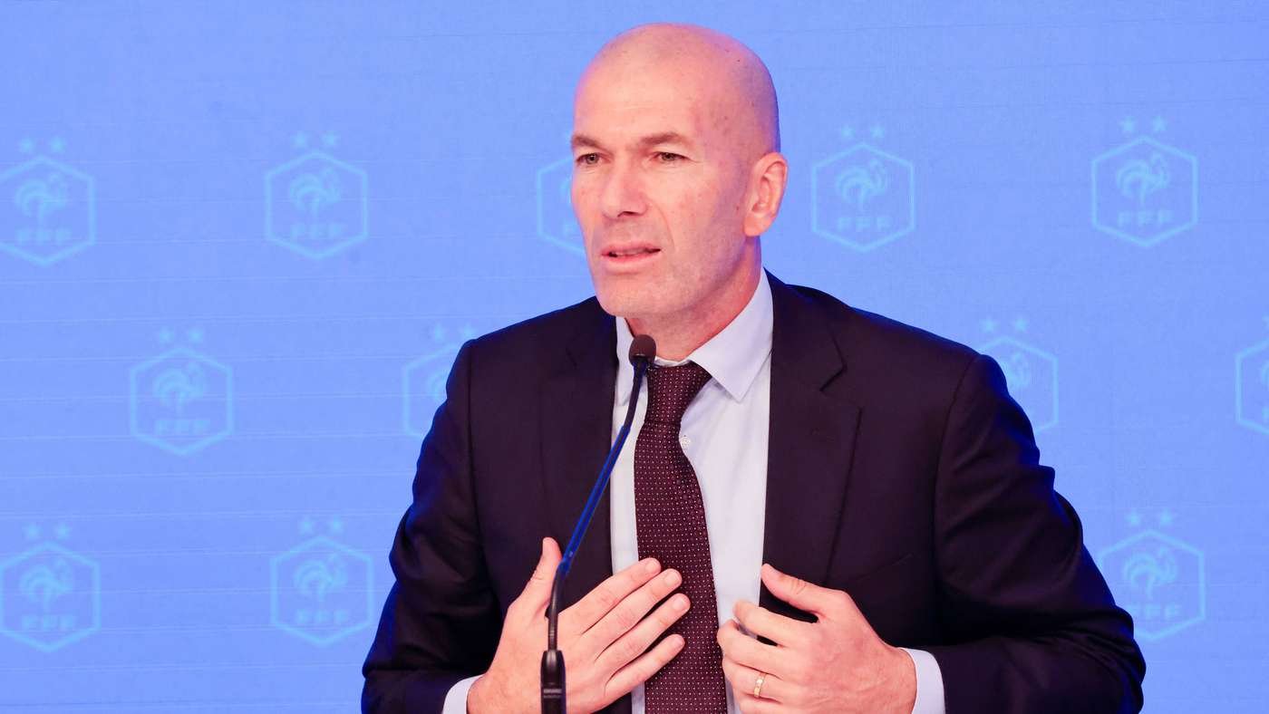 Bald man in a dark suit and red tie speaks into a microphone at a press conference with a blue branded backdrop