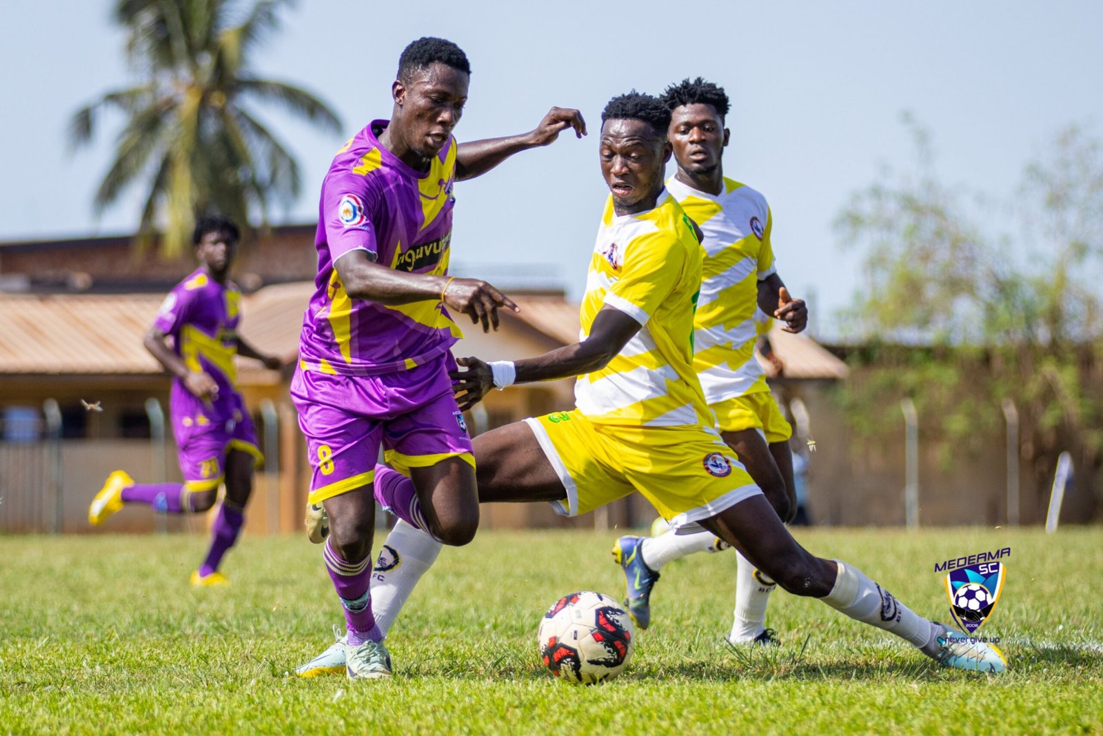 Soccer players in purple and yellow jerseys chase the ball on a grassy field during a match