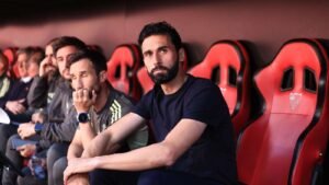 Bearded man in a navy shirt seated on a red dugout bench looking directly at the camera with teammates beside him