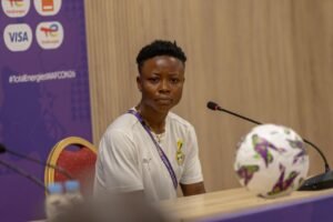Panelist a woman in a white shirt with lanyard sits at a conference table with a microphone in front of her and a decorative ball nearby on the table