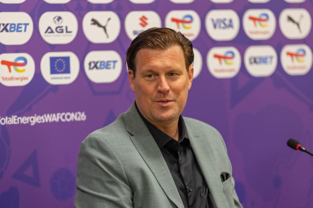 Man in a gray blazer and black shirt sits at a press conference backdrop with sponsor logos behind him
