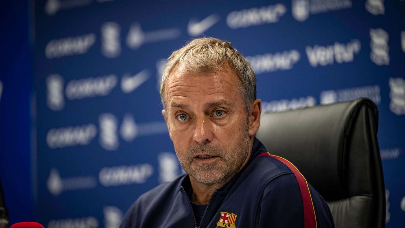 Man with gray hair in an FC Barcelona jacket sits at a press conference blue sponsor wall behind him
