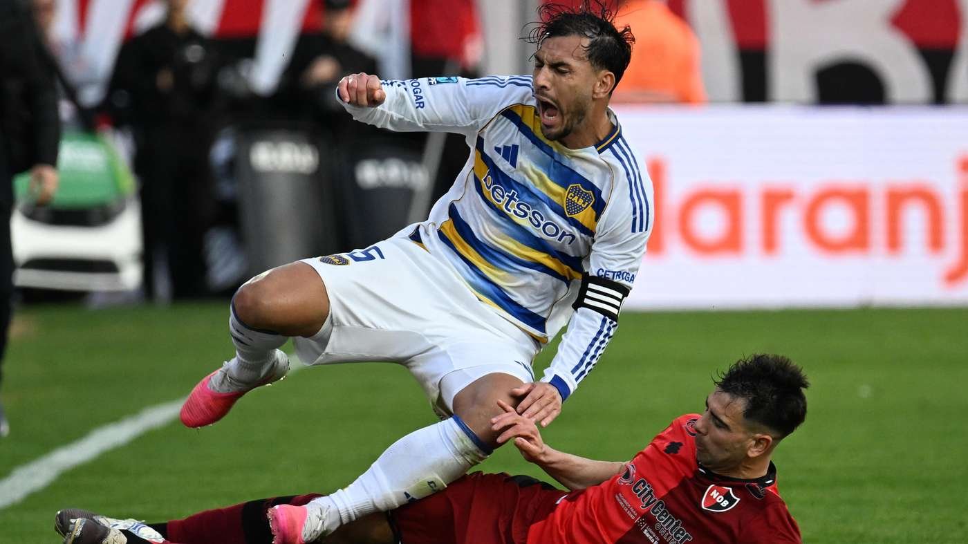 Soccer player in white and blue jersey leaps over a red clad opponent during a tackle on a green field