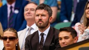 Bearded man in a dark suit and white shirt sits among spectators looking serious at an event