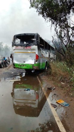 Damaged bus off a rural road into a large puddle with smoke in the background and bystanders nearby