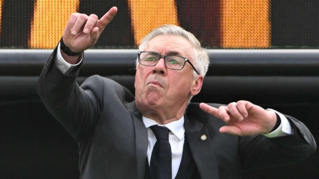 Older man in a dark suit and glasses making a dramatic gesture with one arm raised at a sports venue