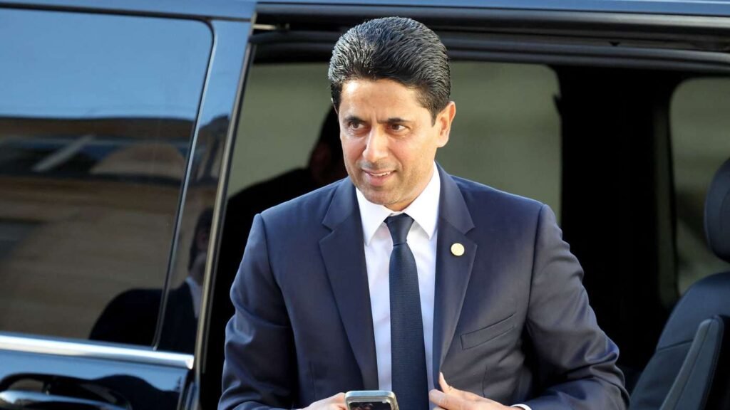 Man in a navy suit exits a black SUV, holding a smartphone and wearing a lapel pin.