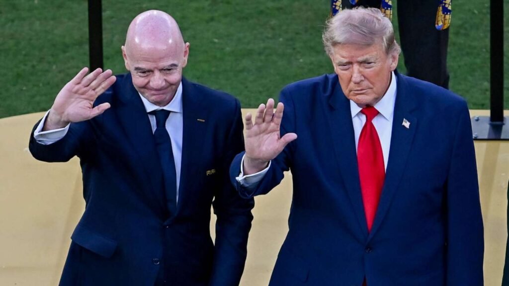 Two men in dark suits wave goodbye on a platform, one bald and the other with a red tie, against a green lawn behind them.