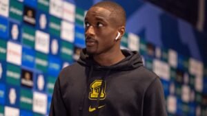 Athlete in a black hoodie with a yellow crest wearing AirPods standing in front of a sponsor backdrop at a press conference