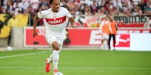 Soccer player in white kit dribbling with a ball on a green field during a match crowd in the stands behind him