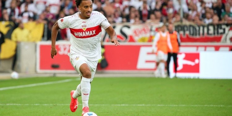 Soccer player in white kit dribbling with a ball on a green field during a match crowd in the stands behind him