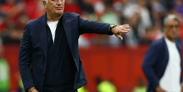 Man in a dark jacket and white shirt gesturing with his arm extended on a sports field stadium stands blurred in the background