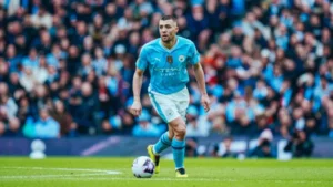 Soccer player in light blue Manchester City kit dribbling a ball on the field with fans in the stands behind him