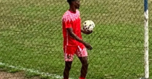 Soccer player in a red uniform standing near a chain link fence controlling a soccer ball at the field edge