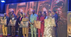 Group of diverse people on a stage at an awards event each holding framed posters while a large portrait filled backdrop looms behind and sparks fly at the side edge of the stage
