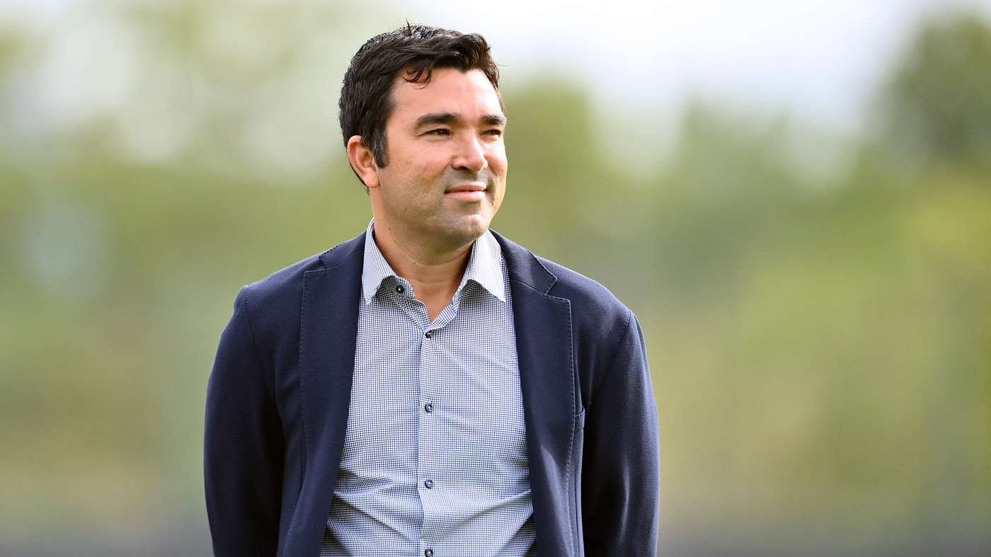 Man in a navy blazer and light checkered shirt standing outdoors on a sunny day in a park like setting