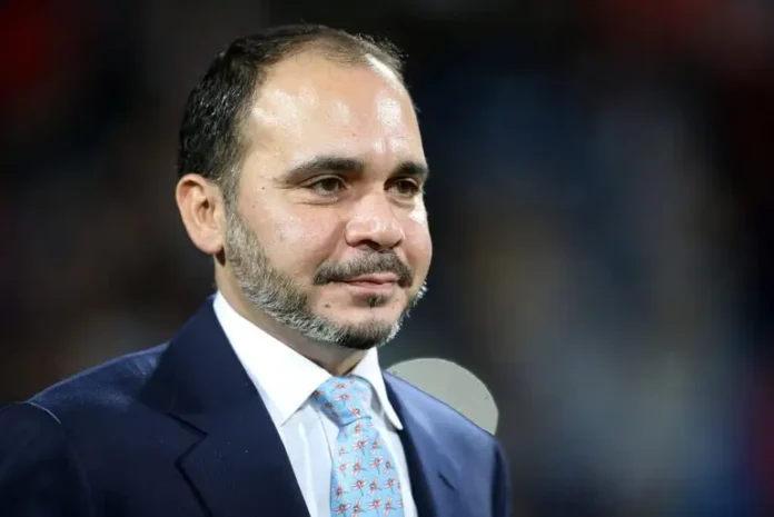 Man in a navy suit and light blue patterned tie speaking at a formal event facing slightly to the right